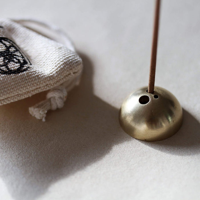 Brass incense holder with a stick on a light surface next to a white hemp bag.