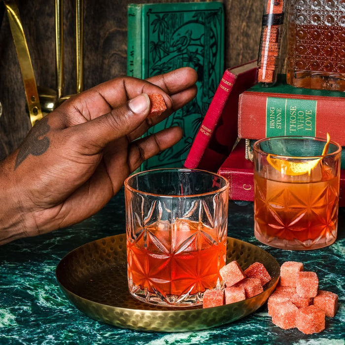 Two glasses of red cocktails on a green table with books and a hand holding a cocktail sugar cube.