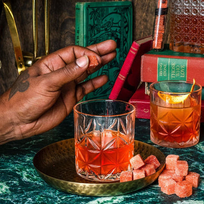Two glasses of red cocktails on a green table with books and a hand holding a cocktail sugar cube.