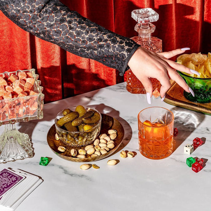 Person holding a sugar cube over a glass with an orange cocktail on a table with snacks and dice.